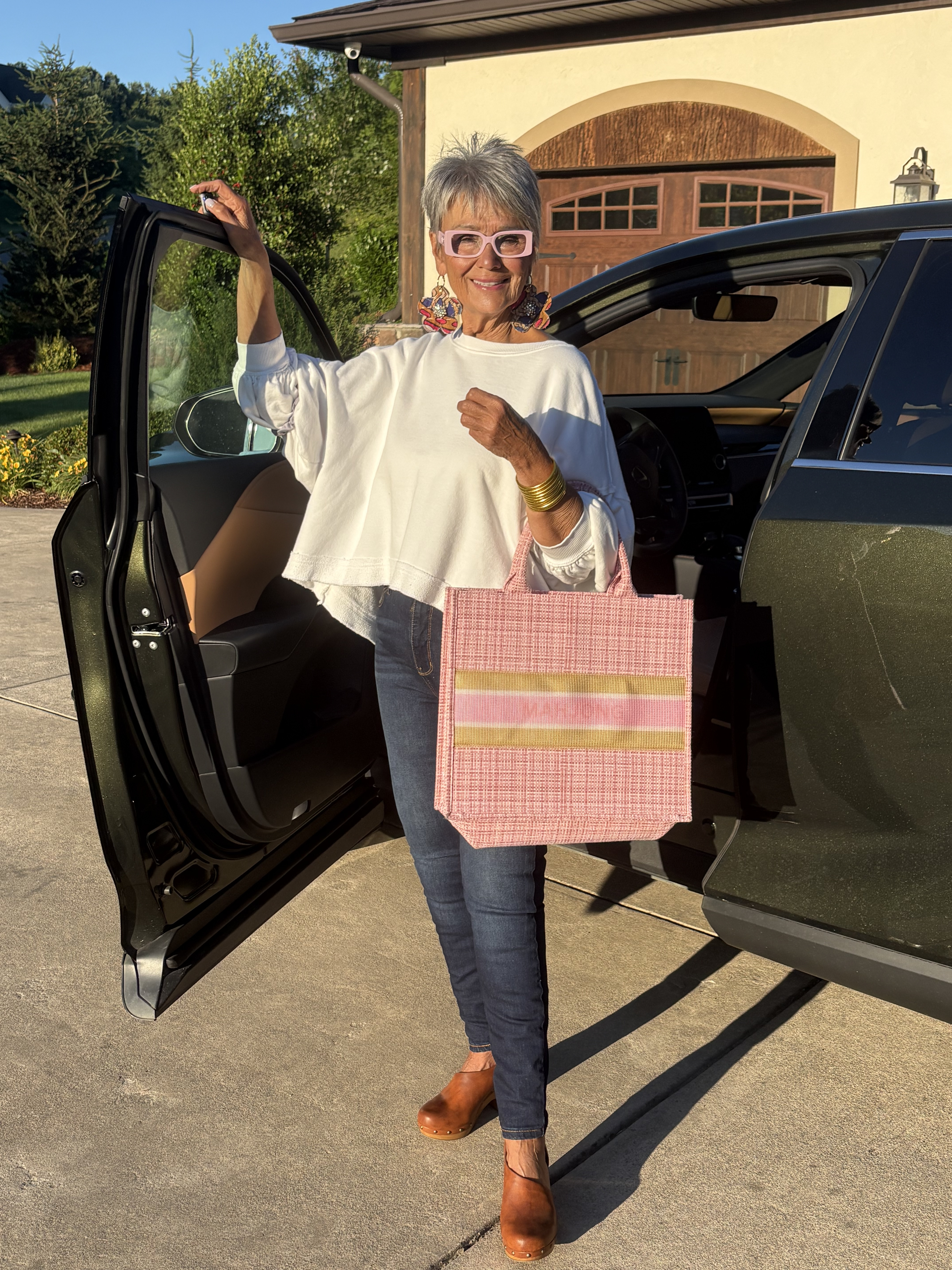Mindy standing by her car holding a pink tote bag, wearing a white top and statement earrings