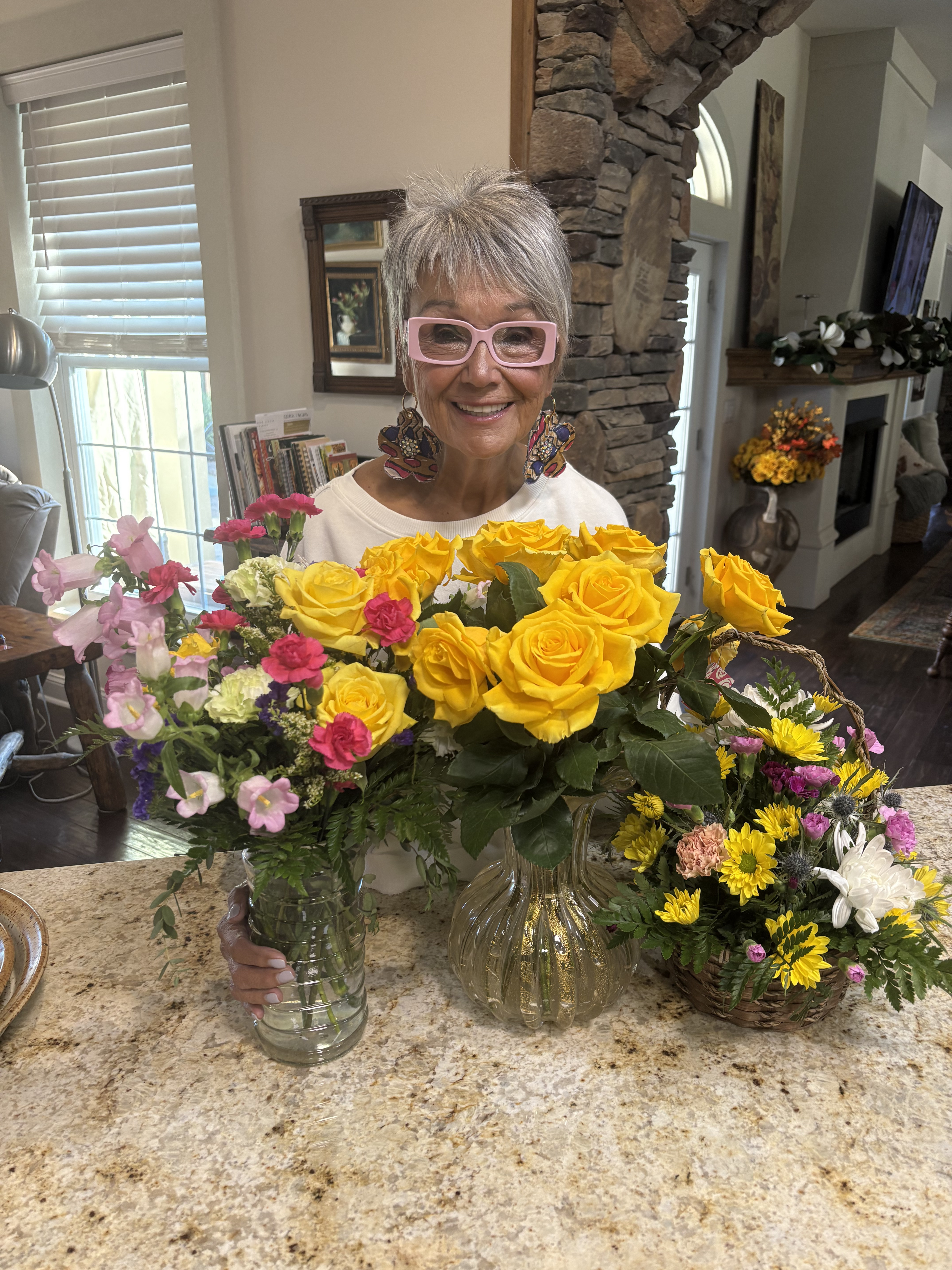 Mindy smiling warmly surrounded by beautiful bouquets of yellow roses and colorful flowers